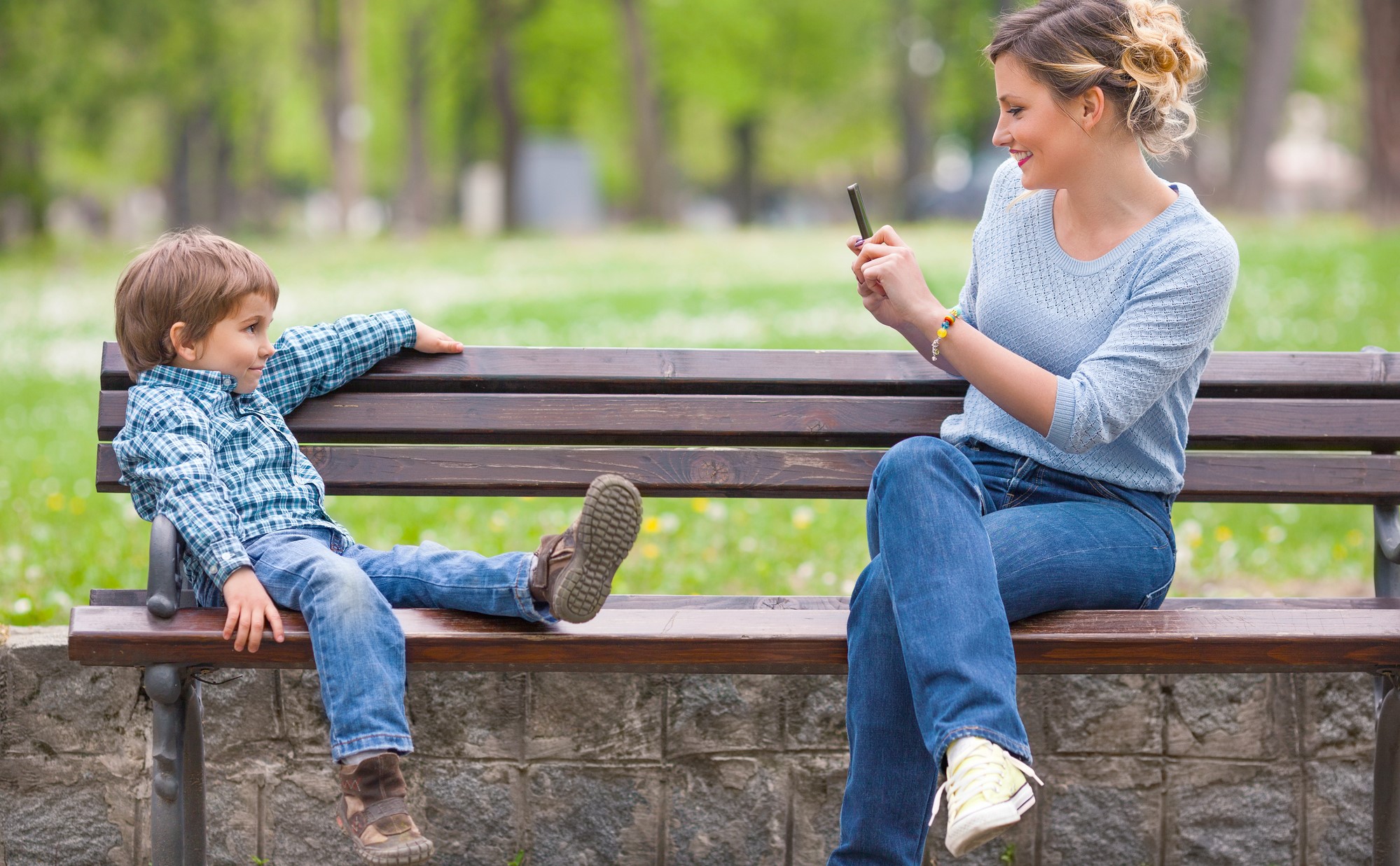 Young mother photographing her cute little son posing on a park bench